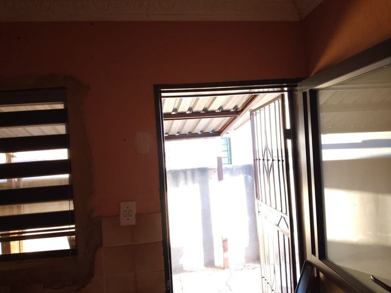 An interior entry area featuring a security gate, aluminium door, and roller blinds, showing signs of unfinished wall plastering around the window frame.