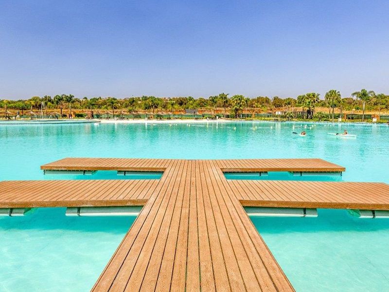 A large, clear swimming lagoon featuring a central wooden jetty and landscaped surroundings, offering a resort-style outdoor recreational space.