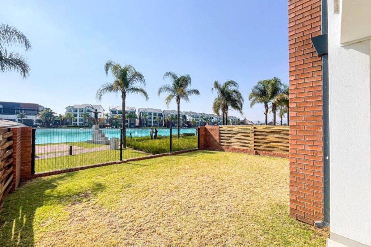 A private rear garden area featuring a lawn, brick walling, and perimeter fencing, with views towards a communal lagoon and residential complex.