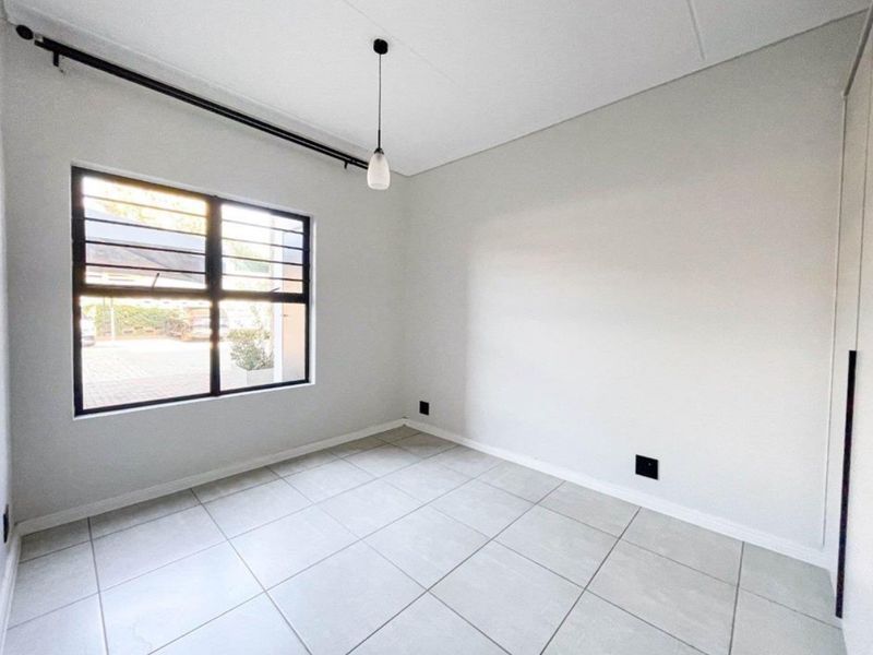 A bright, empty bedroom featuring neutral tiled flooring, a modern pendant light, and a large window with black framing.