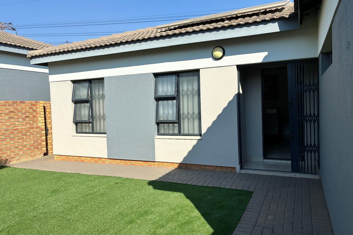 Rear exterior view of the townhouse featuring a paved walkway, artificial lawn, and secure entrance with a metal security gate.