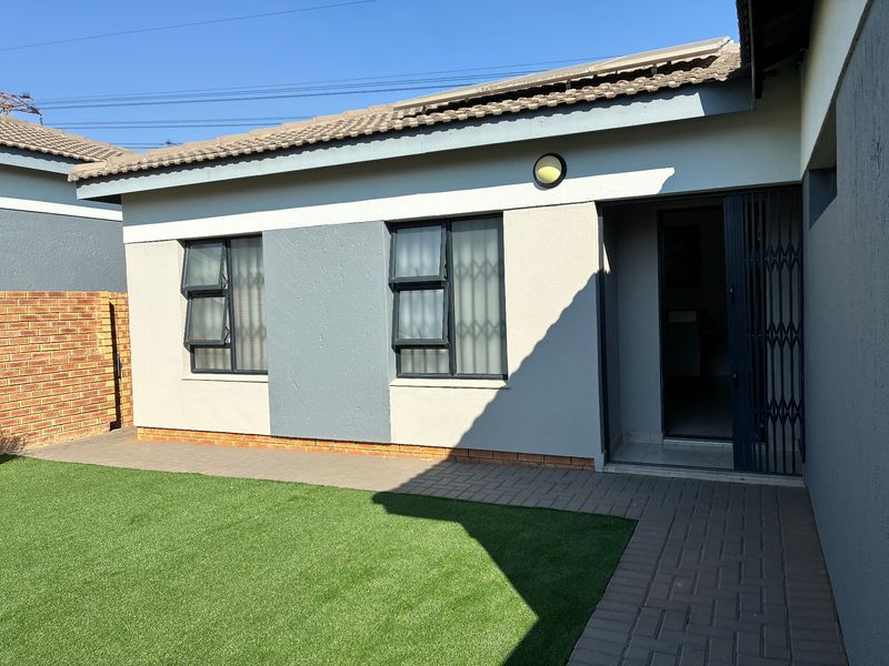 Rear exterior view of the townhouse featuring a paved walkway, artificial lawn, and secure entrance with a metal security gate.