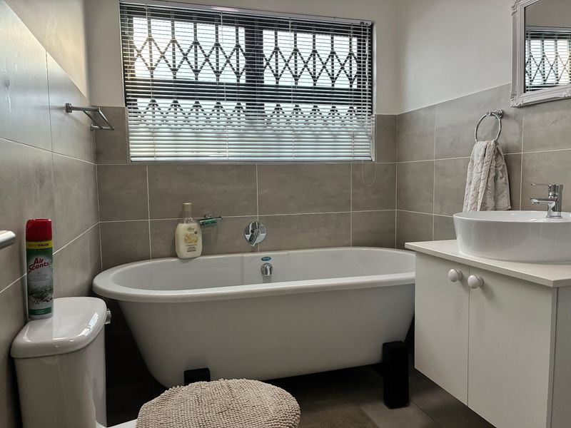 A modern bathroom featuring a freestanding bathtub, a vanity with a vessel sink, and neutral wall tiling with a security-gated window.