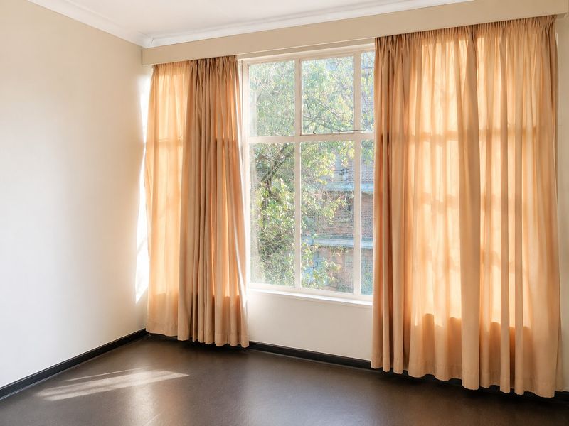 A bright, empty bedroom featuring large multi-pane windows with curtains, neutral walls, and dark flooring, offering a clean canvas for personalization.