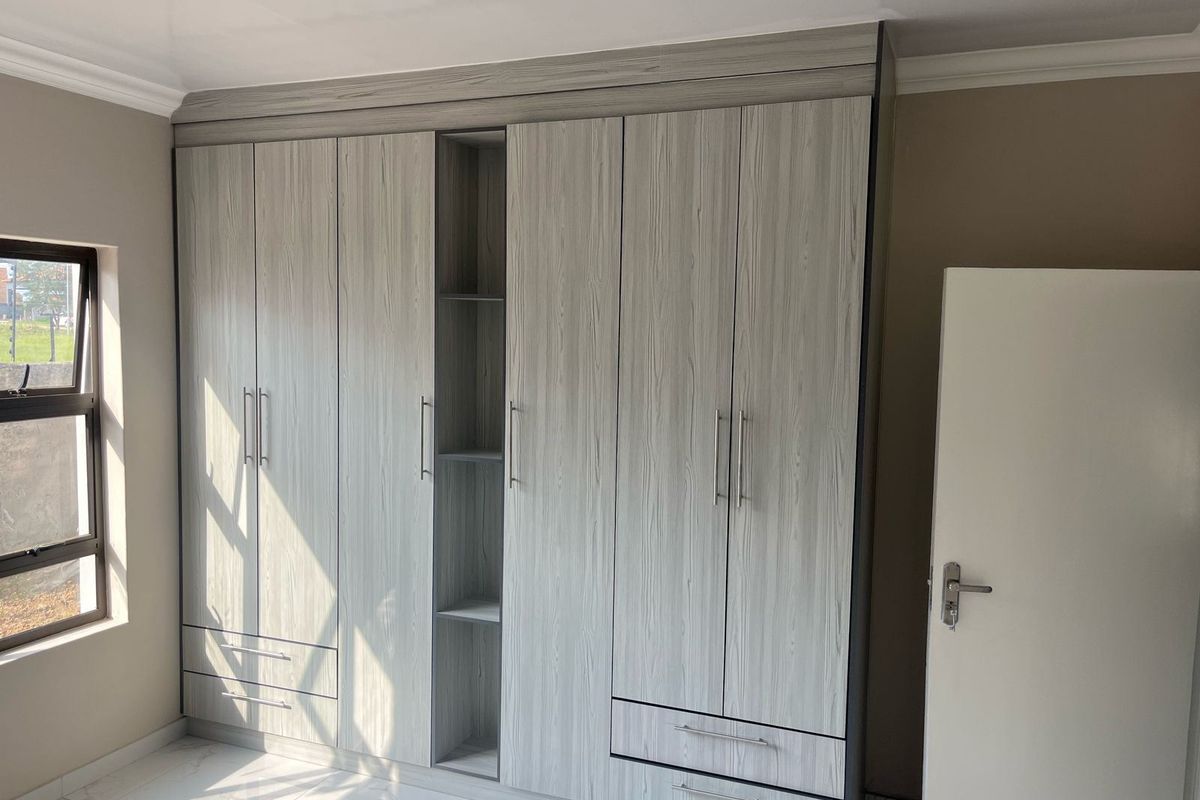 A bright bedroom featuring modern, light wood-grain built-in cupboards with integrated shelving and drawers, complemented by clean tiled flooring.