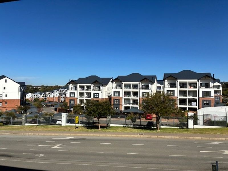 A street-level view of a modern residential apartment complex featuring multi-story buildings with balconies and secure perimeter fencing.