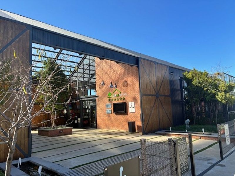Modern industrial-style building facade featuring exposed brickwork, large sliding timber doors, a glass roof section, and a landscaped paved entrance area.