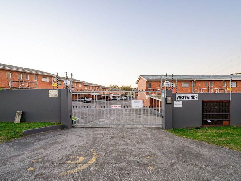 Front entrance view of Westwinds apartment complex featuring a secure gated access point, perimeter wall with electric fencing, and communal post boxes.