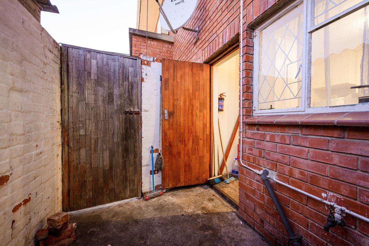 A rear exterior utility area featuring exposed brickwork, a timber door, and concrete flooring, providing access to the apartment's rear entrance.