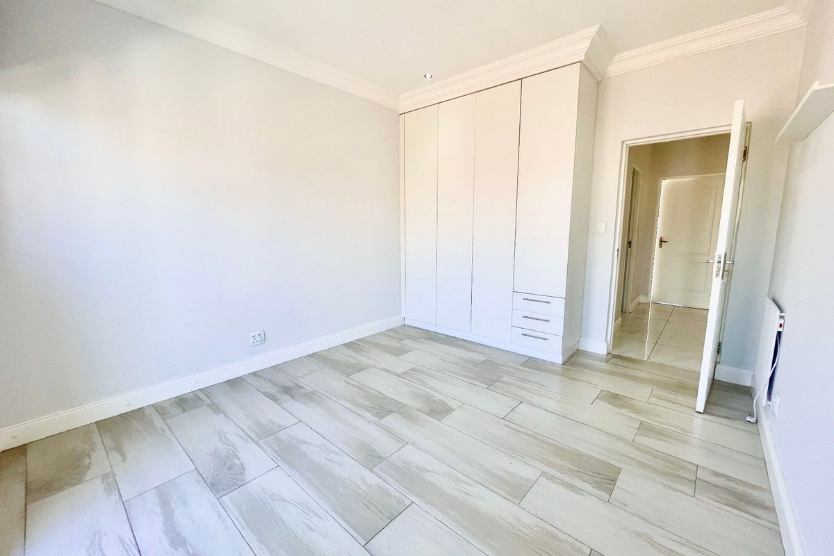 A bright, empty bedroom featuring modern wood-look tiled flooring, built-in cupboards, and recessed downlighting.