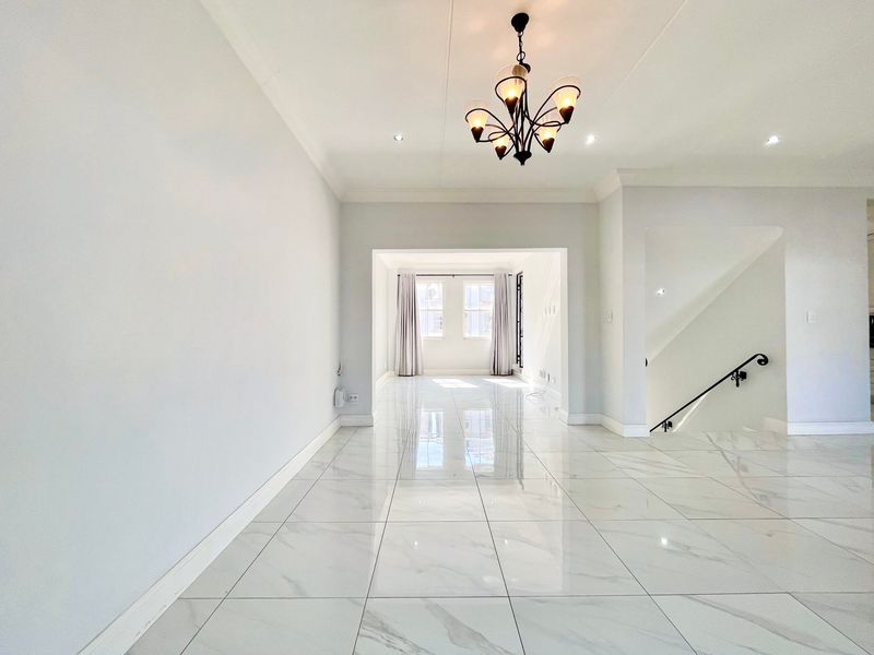 A bright, spacious, and empty living area featuring polished marble-look floor tiles, a decorative chandelier, and a staircase with a wrought iron railing.