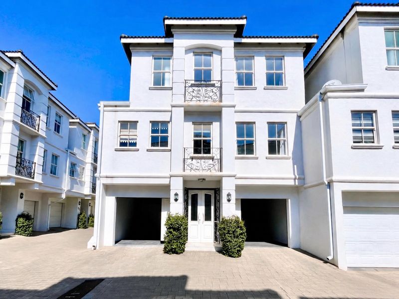 A well-maintained three-story townhouse facade featuring a double garage, paved driveway, and decorative wrought iron balcony railings under a clear blue sky.