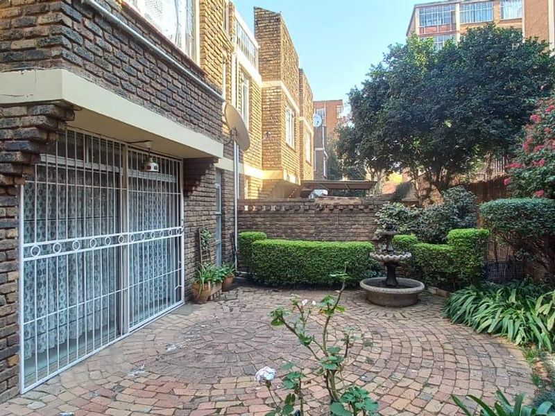 A private rear courtyard featuring a circular brick-paved patio, established garden beds, and a decorative water feature against a classic brick townhouse facade.