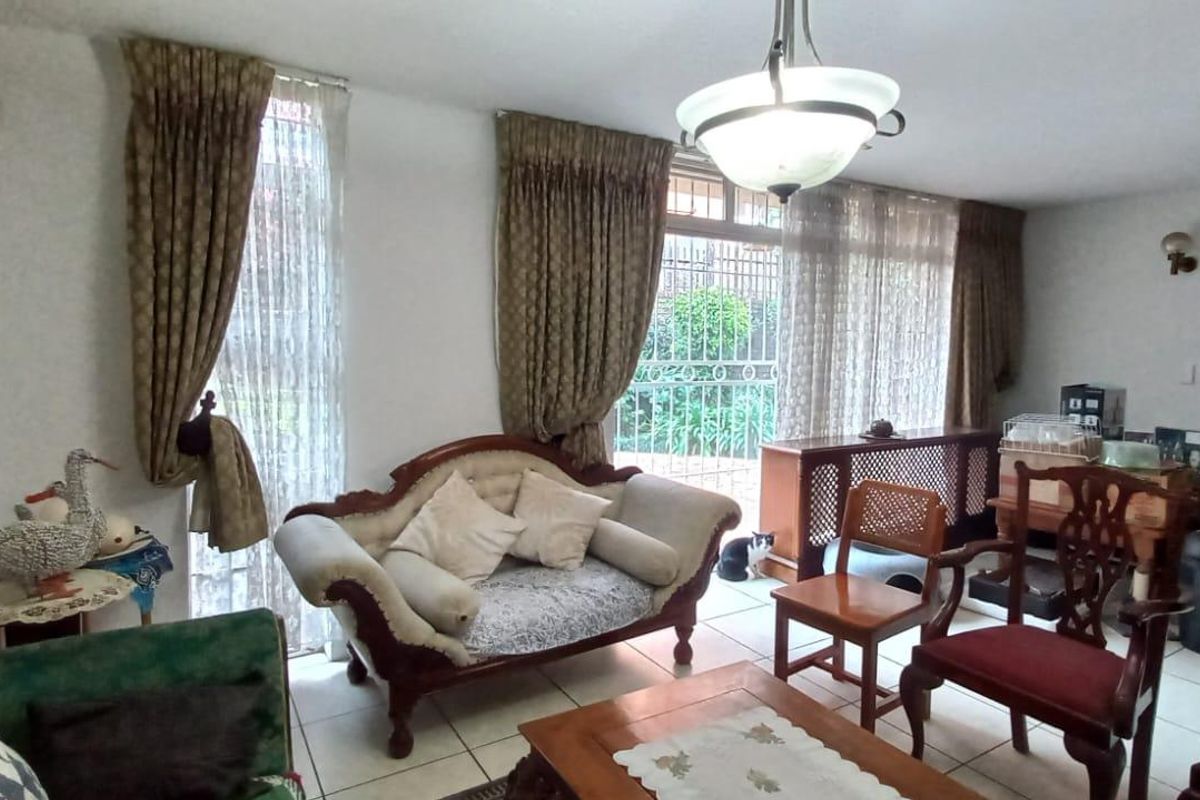 A living room area featuring tiled flooring, large windows with curtains, and access to an outdoor area through a security gate.