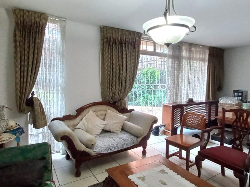 A living room area featuring tiled flooring, large windows with curtains, and access to an outdoor area through a security gate.