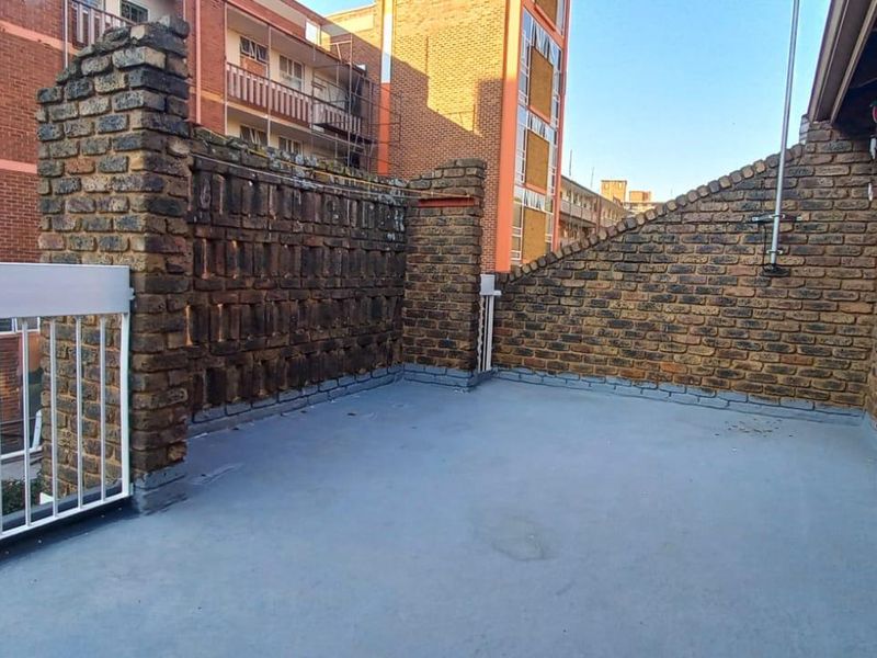 An open-air balcony featuring exposed brick walls, a metal safety railing, and a concrete floor, offering a functional outdoor space.