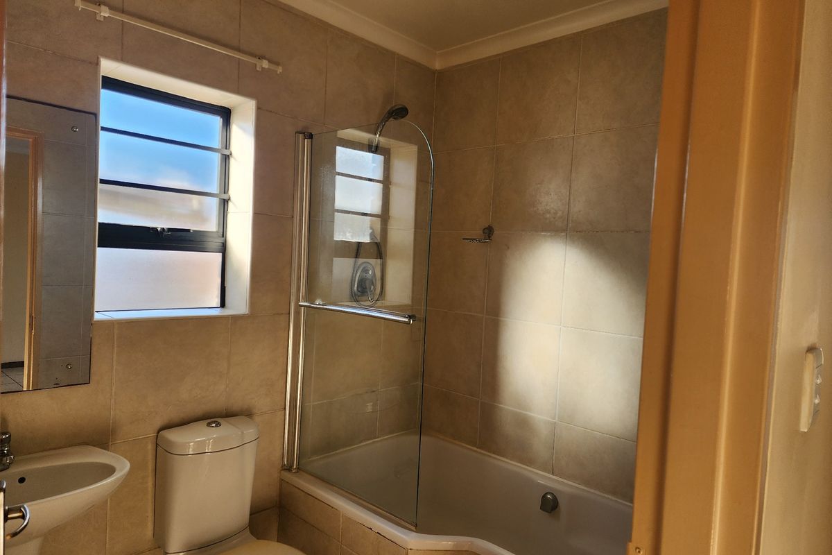 A functional bathroom featuring a bathtub with a glass shower screen, pedestal basin, toilet, and neutral wall tiling.