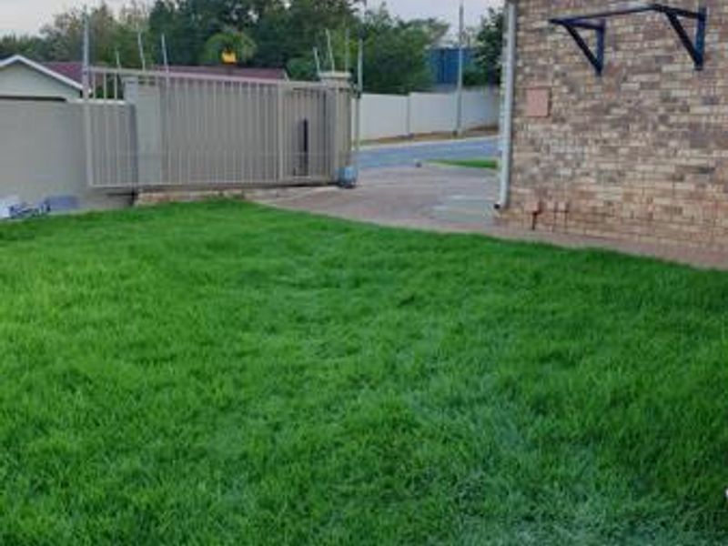 A view of the rear garden featuring a well-maintained lawn, brick exterior wall, and secure perimeter fencing with a sliding gate.