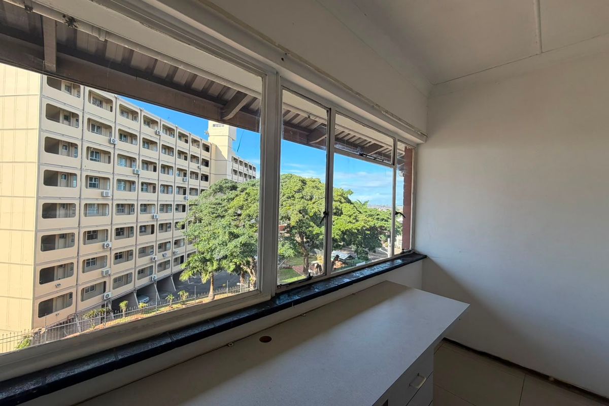 A bright, empty bedroom featuring a built-in desk, tiled flooring, and large windows offering an urban view of the surrounding apartment complex.