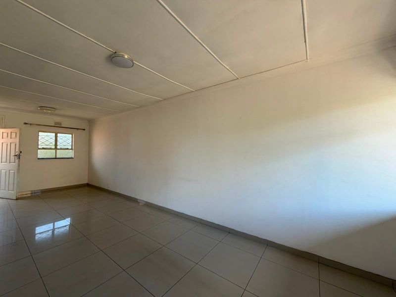 A spacious, empty living area featuring neutral tiled flooring, large windows with security bars, and a simple ceiling design, ready for personalization.