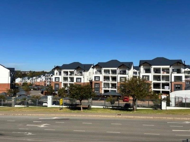 A street-level view of a modern apartment complex featuring multi-story residential blocks, covered parking, and secure perimeter fencing.
