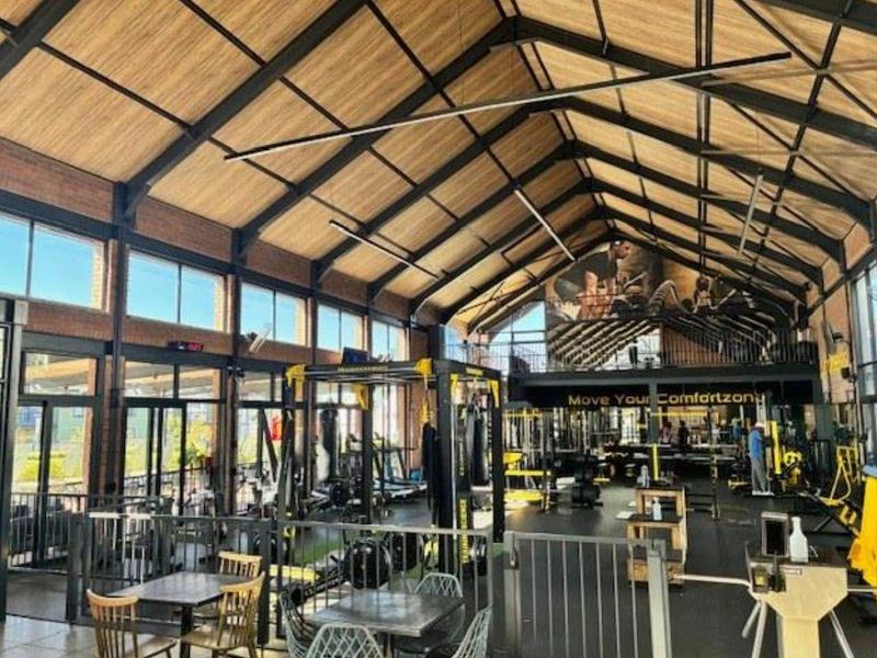 Spacious industrial-style gym featuring high vaulted timber ceilings, exposed steel trusses, large windows, and a mezzanine level with modern fitness infrastructure.