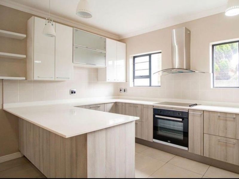 Modern kitchen featuring light wood-grain cabinetry, white stone countertops, built-in oven and hob, stainless steel extractor fan, and neutral tiled finishes.