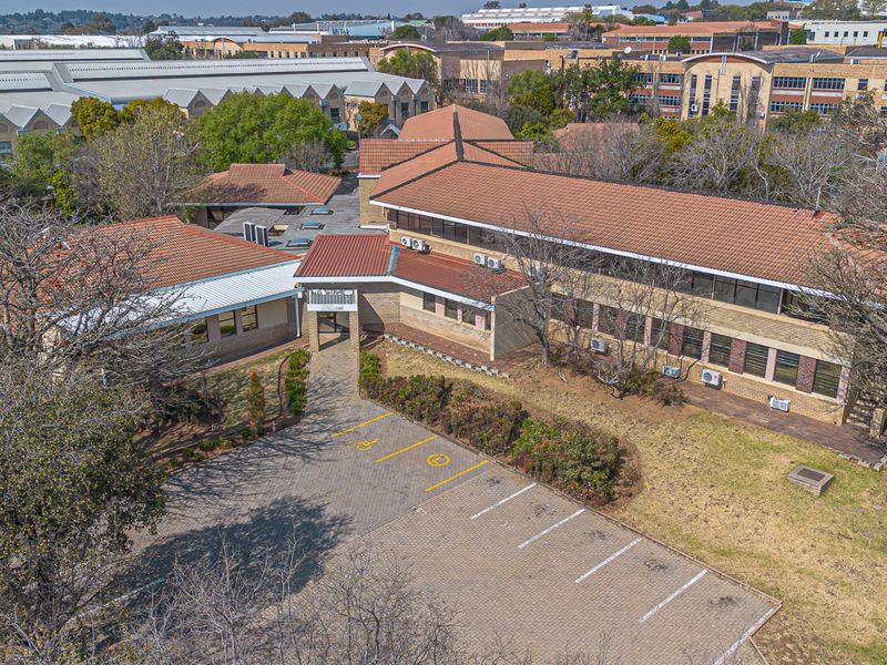 Aerial view of a commercial property featuring a multi-wing brick building with tiled roofing, surrounding landscaped areas, and a paved parking lot.