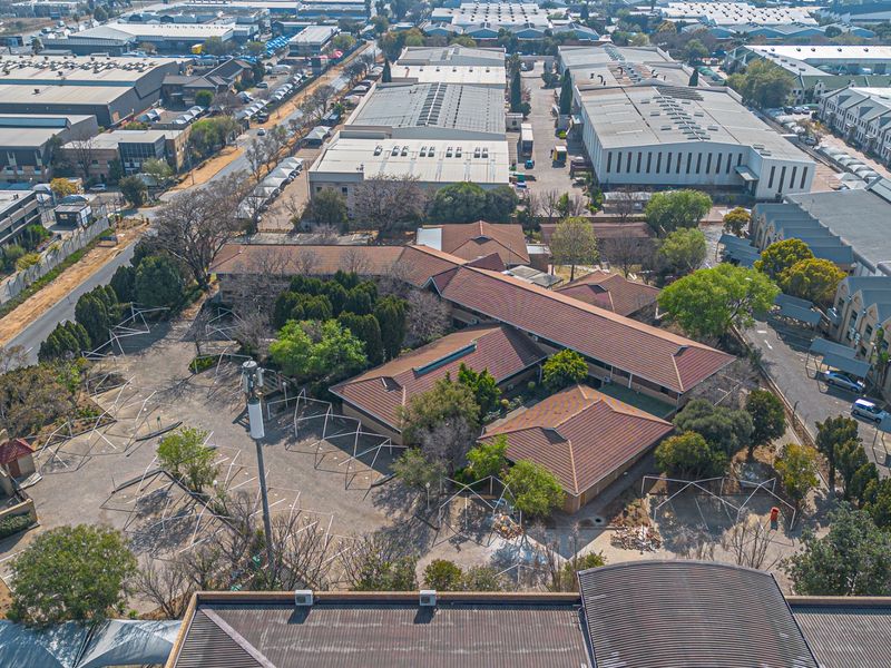 Aerial view of a commercial property complex featuring multiple buildings with tiled roofs, extensive paved parking areas, and established landscaping within an industrial park setting.