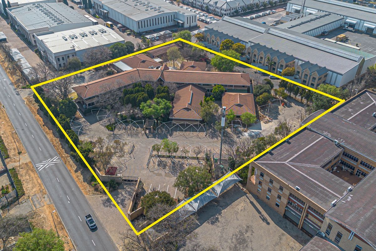 Aerial view of a commercial property complex featuring multiple buildings, a large paved parking area, and established landscaping within a fenced perimeter.