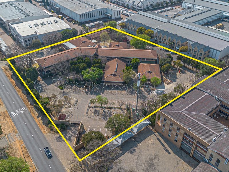 Aerial view of a commercial property complex featuring multiple buildings, a large paved parking area, and established landscaping within a fenced perimeter.
