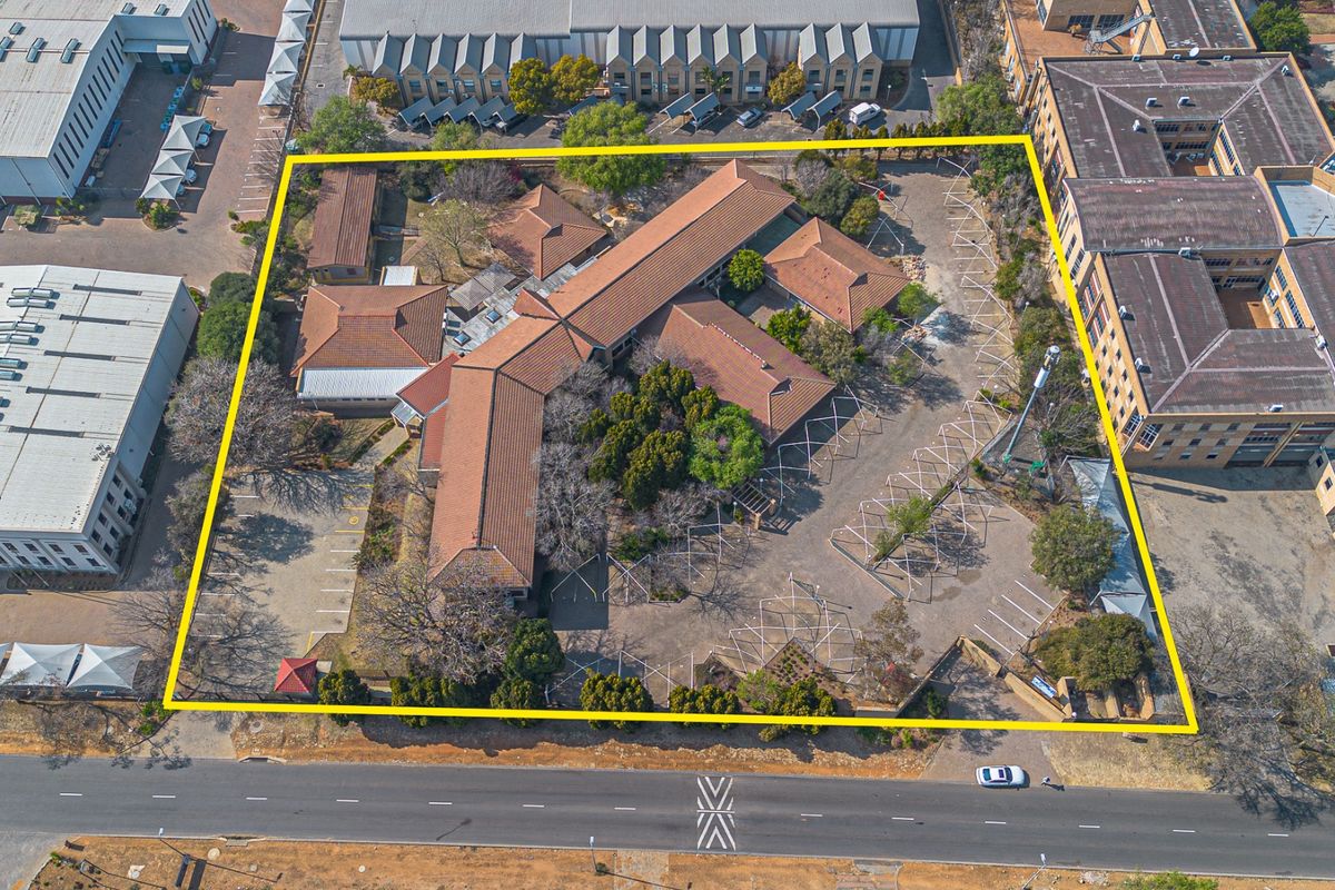 Aerial view of a commercial property complex featuring multiple buildings with tiled roofs, extensive paved parking, and landscaped grounds within a defined perimeter.