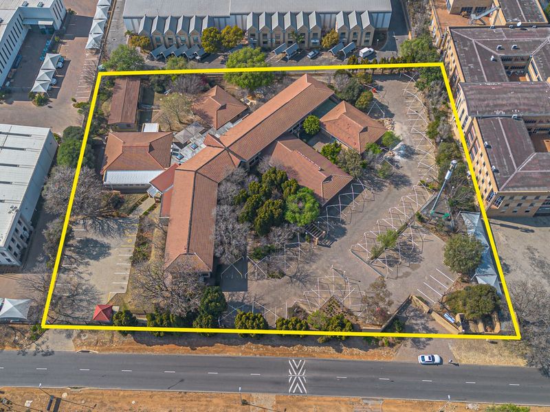 Aerial view of a commercial property complex featuring multiple buildings with tiled roofs, extensive paved parking, and landscaped grounds within a defined perimeter.