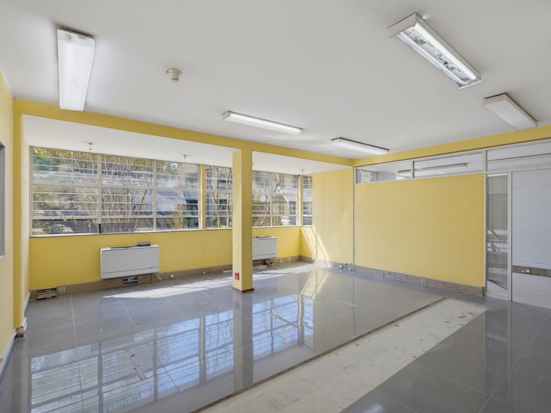 An empty commercial office space featuring large windows, tiled flooring, and fluorescent lighting, currently requiring minor floor repairs.