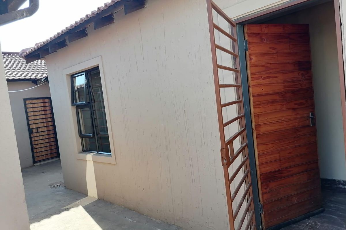 Exterior view of the rear building facade featuring a wooden door with a security gate, aluminium window, and concrete paved walkway.