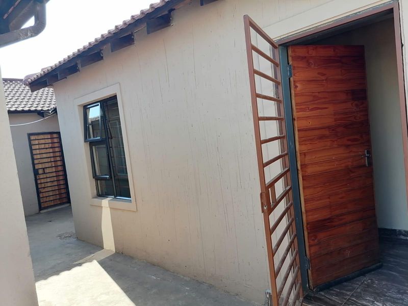 Exterior view of the rear building facade featuring a wooden door with a security gate, aluminium window, and concrete paved walkway.