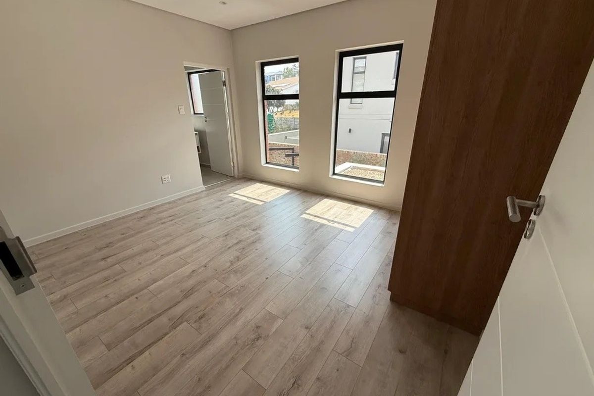 A bright, empty bedroom featuring modern laminate flooring, built-in cupboards, and large aluminium-framed windows allowing for ample natural light.