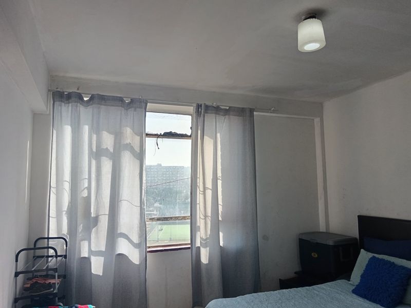 A bedroom featuring a window with curtains and a ceiling light fixture, showing signs of wear on the walls and window frame.