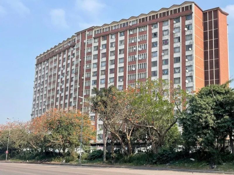 A multi-story apartment building exterior featuring a brick facade and numerous balconies, viewed from the adjacent street.