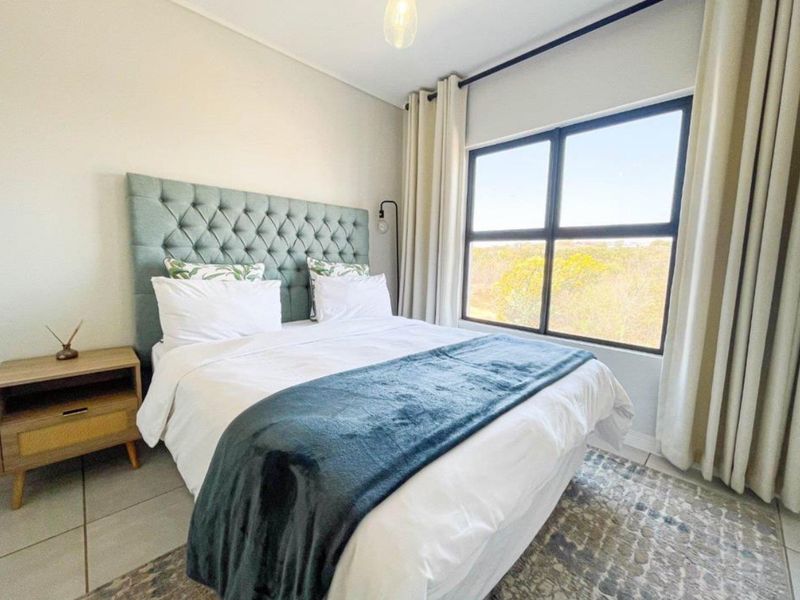 A well-maintained bedroom featuring tiled flooring, neutral walls, and a large window with black aluminium frames offering natural light and scenic views.