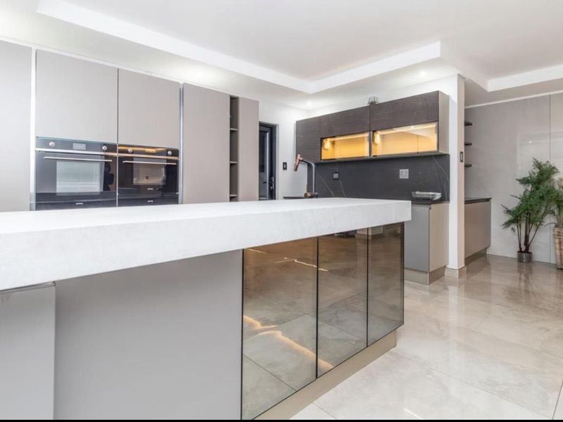 A modern, high-end kitchen featuring a large central island, sleek cabinetry, built-in double ovens, and polished tiled flooring with integrated lighting.