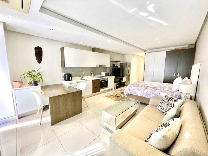 A modern open-plan studio apartment featuring a sleek kitchen with built-in cabinetry, tiled flooring, and integrated storage solutions in a well-maintained living space.