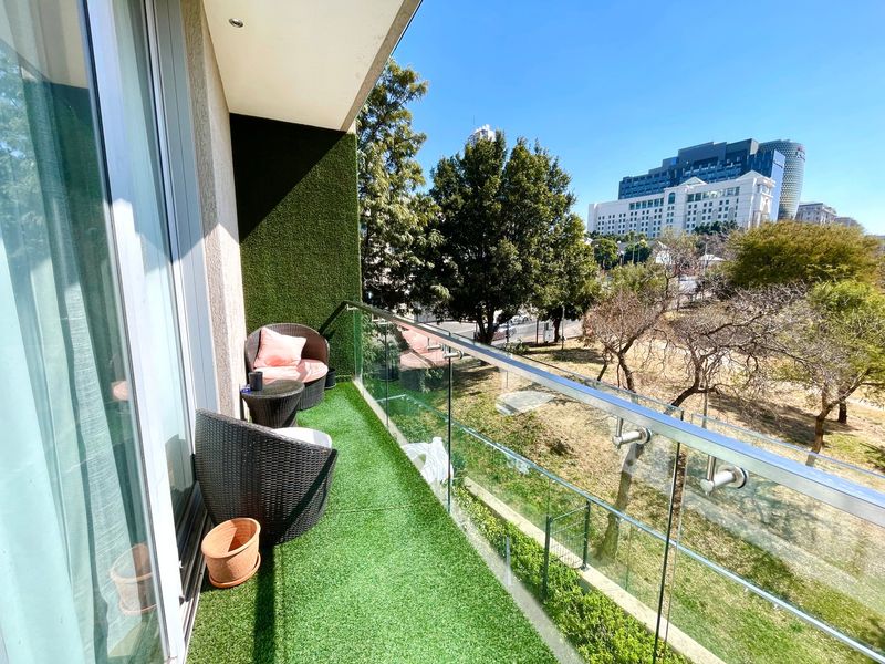 A modern balcony featuring a glass balustrade with stainless steel handrails, artificial grass flooring, and recessed lighting, offering an elevated view of the surrounding area.
