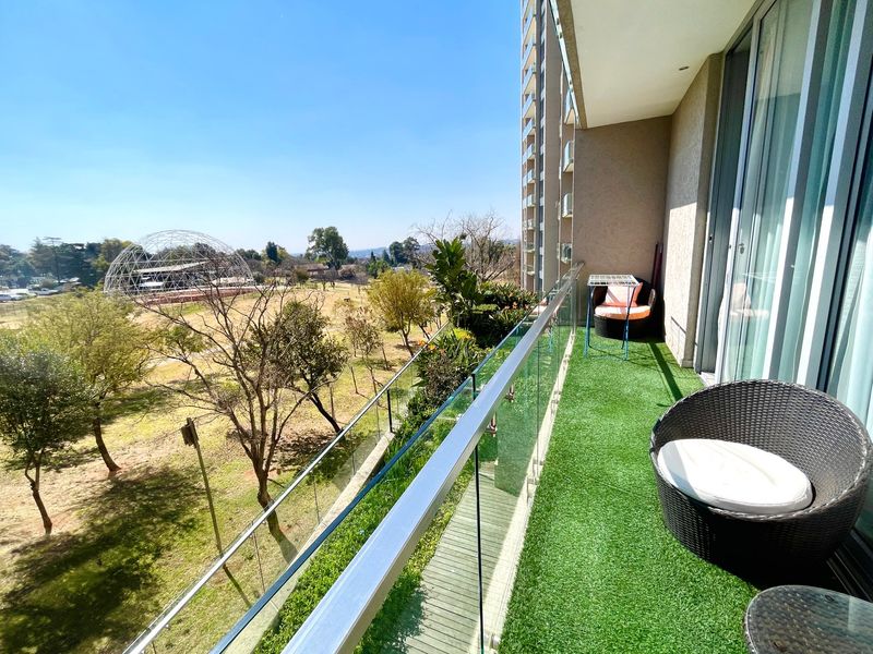 A modern apartment balcony featuring a glass balustrade and artificial turf, offering expansive views over the surrounding parkland and neighborhood.
