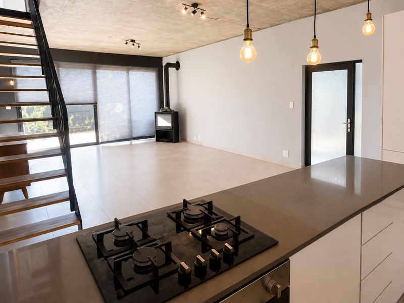 Modern open-plan living area featuring a sleek kitchen with gas hob, exposed concrete ceilings, and a striking open-tread staircase leading to the upper level.