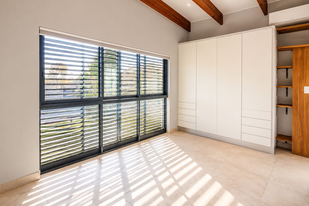 A bright, modern bedroom featuring tiled floors, exposed timber ceiling beams, built-in cabinetry, and large windows with integrated external louvre shutters.