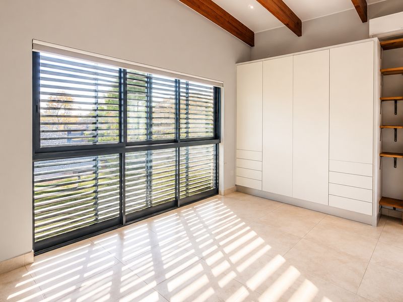 A bright, modern bedroom featuring tiled floors, exposed timber ceiling beams, built-in cabinetry, and large windows with integrated external louvre shutters.