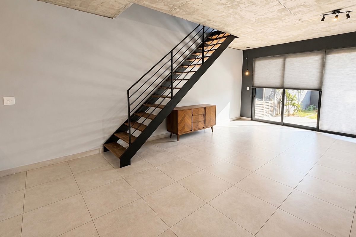 Spacious open-plan living area featuring modern exposed concrete ceilings, sleek metal staircase, large sliding glass doors, and neutral tiled flooring.