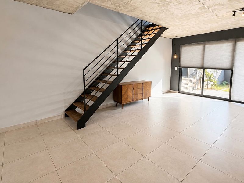 Spacious open-plan living area featuring modern exposed concrete ceilings, sleek metal staircase, large sliding glass doors, and neutral tiled flooring.