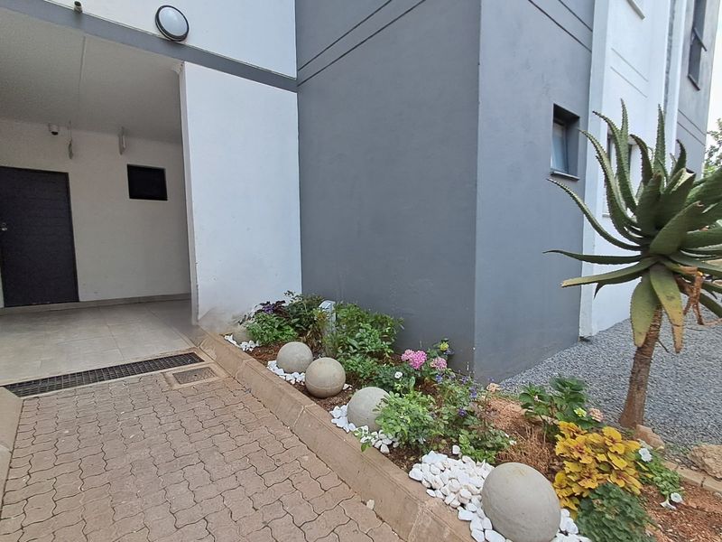 A clean exterior view of the apartment entrance featuring a paved walkway, modern grey and white facade, and a well-maintained landscaped garden bed.